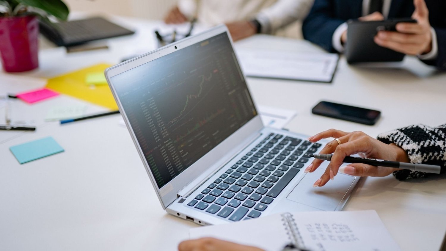 Person using a laptop displaying financial charts during a team meeting, with colleagues, notes and a phone on the table.