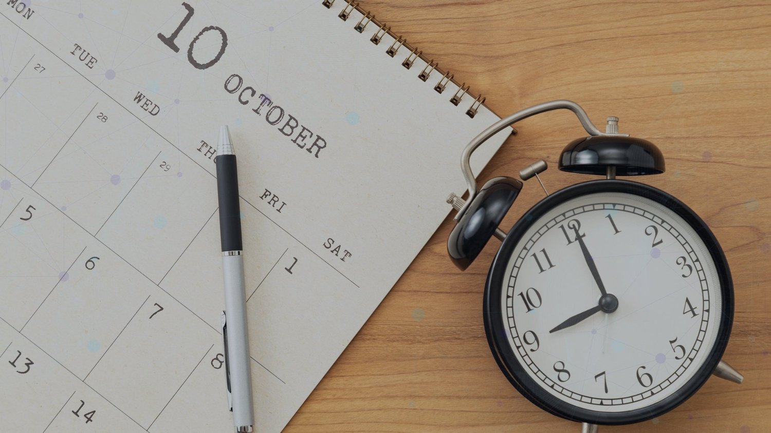 Alarm clock beside a calendar and pen on a wooden desk.